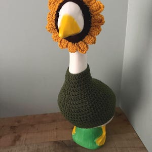 May include: A white ceramic goose figurine wearing a dark olive green crocheted sweater and a yellow and brown crocheted sunflower headpiece that frames the goose's face. The goose has a bright yellow beak and stands on a green base.