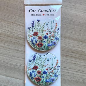 May include: Two car coasters with a floral design featuring red, blue, and purple flowers. The coasters are packaged in a clear plastic sleeve. The text "Car Coasters Handmade with love" is printed on the packaging.