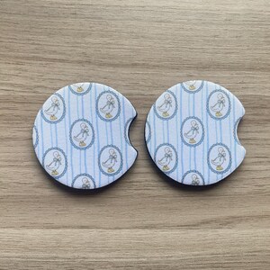 May include: Two round car coasters with a duck pattern. The coasters have a blue and white striped background with oval medallions featuring a duck design. The coasters are made of a absorbent material and have a small notch for easy removal.