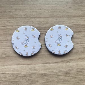 May include: Two white car coasters with a duck and floral pattern. Each coaster features a duck wearing a blue bow and yellow boots, surrounded by small flowers and duck silhouettes. The coasters have a curved cutout for easy removal.