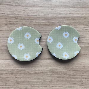 May include: Two round car coasters with a daisy pattern. The coasters have a light green background with a grid pattern and white daisies with yellow centers. Each coaster has a curved cutout for easy removal.