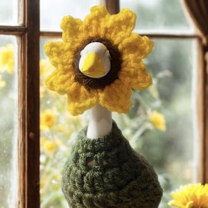 Puede incluir: Una caprichosa figura de pato de cerámica con cabeza de girasol y cuerpo tejido en verde. El pato tiene cuello blanco y pico amarillo. La figura se exhibe sobre una superficie de madera cerca de una ventana con un fondo borroso de flores amarillas.
