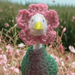 Puede incluir: Un pato de ganchillo hecho a mano, adornado con un sombrero de flores rosa y un cuerpo verde, ambientado en un telón de fondo de flores y follaje rosas. El pato presenta una cara blanca y un pico amarillo, evocando una estética encantadora inspirada en el jardín.