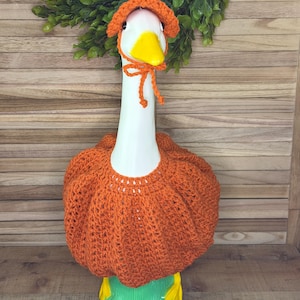 May include: A white ceramic goose figurine wearing a bright orange crocheted pumpkin-shaped outfit that covers its body and a matching orange crocheted bonnet with ties under its yellow beak. The goose has yellow feet and stands upright.