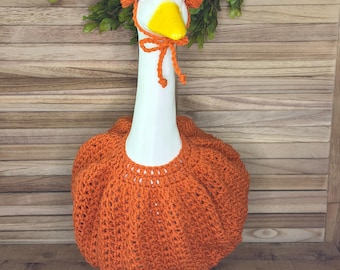 Könnte beinhalten: Eine weiße Gänsefigur aus Keramik, die ein leuchtend orangefarbenes, gehäkeltes kürbisförmiges Outfit trägt, das den Körper bedeckt, sowie eine passende gehäkelte Haube mit Bändern unter dem gelben Schnabel. Die Gans hat gelbe Füße.