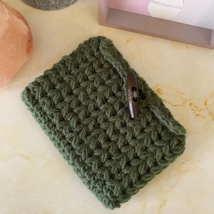 May include: Handmade green crochet phone case with a wooden button closure. The case is made of a textured, woven yarn and is designed to protect a phone. The case is a dark green color.