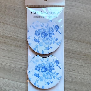 May include: Two car coasters with a white background and a blue floral pattern are displayed in a clear plastic package. The package has the text "Car Coasters" and "Handmade with love".