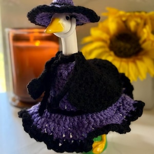 Pu&ograve; includere: Una statuetta di oca bianca che indossa un completo all'uncinetto viola e nero composto da un cappello a punta a tesa larga, un mantello nero e un vestito viola con un orlo nero a balze. L'oca ha un becco giallo brillante e poggia su una base verde.
