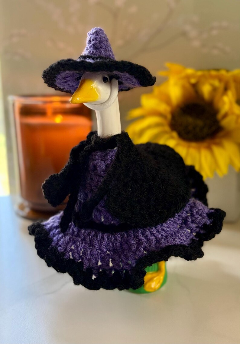 Pu&ograve; includere: Una statuetta di oca bianca che indossa un completo all'uncinetto viola e nero composto da un cappello a punta a tesa larga, un mantello nero e un vestito viola con un orlo nero a balze. L'oca ha un becco giallo brillante e poggia su una base verde.