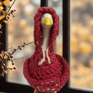 May include: A ceramic goose figurine wearing a red knitted hoodie with white drawstrings. The goose has a white neck, yellow beak, and green and yellow base. The background is a blurred view of a window and foliage.