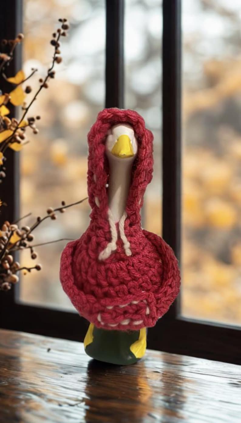May include: A ceramic goose figurine wearing a red knitted hoodie with white drawstrings. The goose has a white neck, yellow beak, and green and yellow base. The background is a blurred view of a window and foliage.