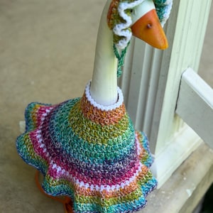 May include: A decorative goose figurine wearing a hand-crocheted outfit consisting of a flared, multi-colored dress and a matching bonnet. The yarn features a variegated pattern of green, purple, pink, and orange, accented with white trim.