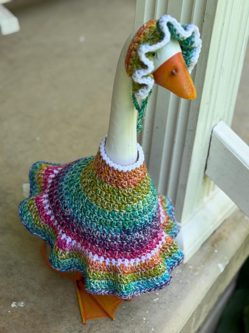 May include: A decorative goose figurine wearing a hand-crocheted outfit consisting of a flared, multi-colored dress and a matching bonnet. The yarn features a variegated pattern of green, purple, pink, and orange, accented with white trim.