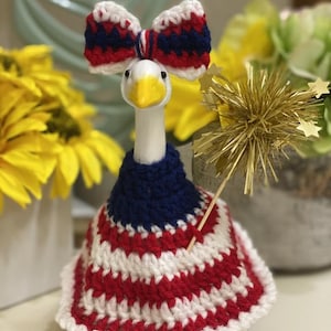 May include: A ceramic goose figurine with a patriotic theme. The goose wears a red, white, and blue crocheted outfit, including a bow and a dress. It holds a gold starburst decoration, perfect for a Fourth of July celebration.