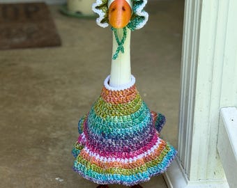 Könnte beinhalten: Eine weiße Gänsefigur, die ein mehrfarbiges Häkelkleid aus meliertem Garn mit gerüschtem Saum und eine passende Häkelhaube mit weißem Bogenkantenabschluss und grünen Bindebändern trägt. Die Gans hat einen leuchtend orangefarbenen Schnabel und orangefarbene Schwimmfüße.