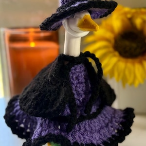 Pu&ograve; includere: Una statuetta di oca bianca che indossa un cappello da strega all'uncinetto viola e nero, un mantello nero e un vestito viola con un orlo nero a balze. L'oca ha un becco giallo ed &egrave; vestita con un set coordinato di capi fatti all'uncinetto.