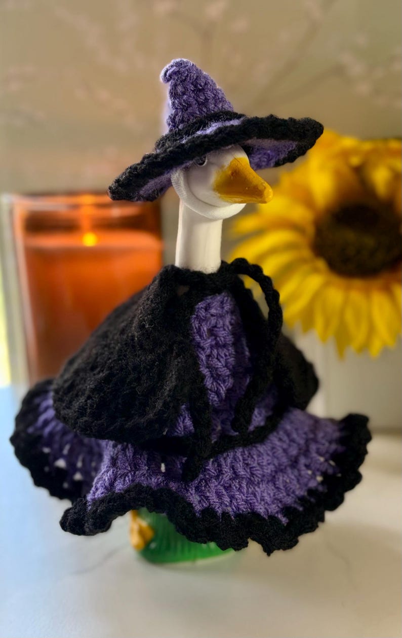 Pu&ograve; includere: Una statuetta di oca bianca che indossa un cappello da strega all'uncinetto viola e nero, un mantello nero e un vestito viola con un orlo nero a balze. L'oca ha un becco giallo ed &egrave; vestita con un set coordinato di capi fatti all'uncinetto.