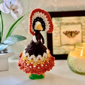 May include: A small white ceramic goose figurine wearing a crocheted outfit. The outfit consists of a multi-layered skirt in dark brown, cream, mustard yellow, and burnt orange, paired with a matching bonnet featuring the same color palette.