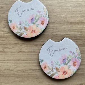 May include: Two car coasters with a floral design in shades of pink, peach, and purple. The name "Emma" is written in cursive on each coaster. The coasters have a black border and a cutout for easy removal.