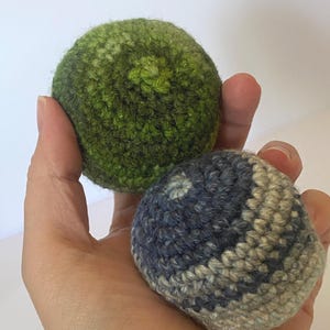 May include: Two spherical crochet objects. One sphere is a solid, textured dark green with a lighter green centre point. The second sphere features a variegated pattern of dark blue, slate grey, and cream-coloured yarn, also with a central point.