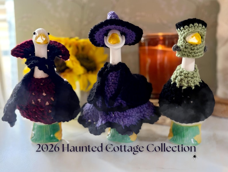 Pu&ograve; includere: Tre statuette di oche in ceramica bianca con abiti all'uncinetto: un vampiro con mantello bordeaux e nero, una strega con vestito e cappello viola, e un mostro con copricapo verde e colletto nero. Testo: 2026 Haunted Cottage Collection.
