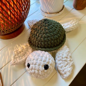 May include: A handmade crochet turtle toy with a green shell, white head, and white flippers. The turtle has black safety eyes and is made of soft, textured yarn. The toy is sitting on a white surface.