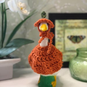 Peut inclure: Une figurine d'oie en céramique blanche portant une tenue en forme de citrouille crochetée à la main. La tenue comprend une housse de corps orange texturée et un bonnet orange assorti avec une petite tige verte sur le dessus, noué sous le menton.