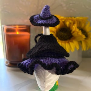 Pu&ograve; includere: Un copribottiglia a forma di cappello da strega all'uncinetto viola e nero, con corona a punta e tesa larga arricciata. Il cappello &egrave; progettato per adattarsi al collo di una bottiglia, con un cappellino abbinato posizionato sul tappo.