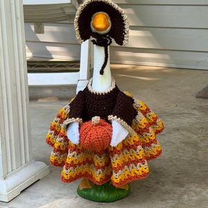 May include: A white goose statue wearing a dark brown crocheted bonnet and a matching dark brown bodice with a multi-coloured skirt featuring horizontal stripes of orange, yellow, and tan yarn. The goose holds a small, round, orange crocheted pumpkin.