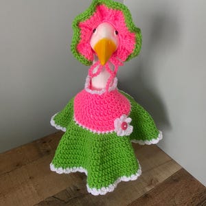 May include: A whimsical goose figurine wearing a handmade crochet outfit. The outfit includes a pink and green dress, a matching bonnet, and a white collar. The goose has a yellow beak and is set against a neutral background.