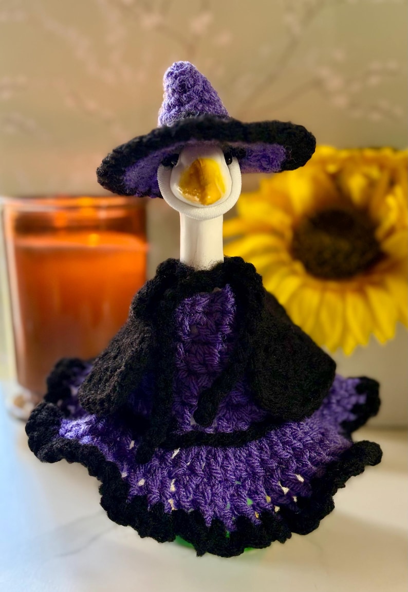 Pu&ograve; includere: Una statuetta di oca in ceramica bianca che indossa un completo lavorato all'uncinetto a mano in viola e nero. L'insieme include un cappello a punta, una piccola mantella e una gonna a balze, tutto con una trama in lana e bordi scuri.