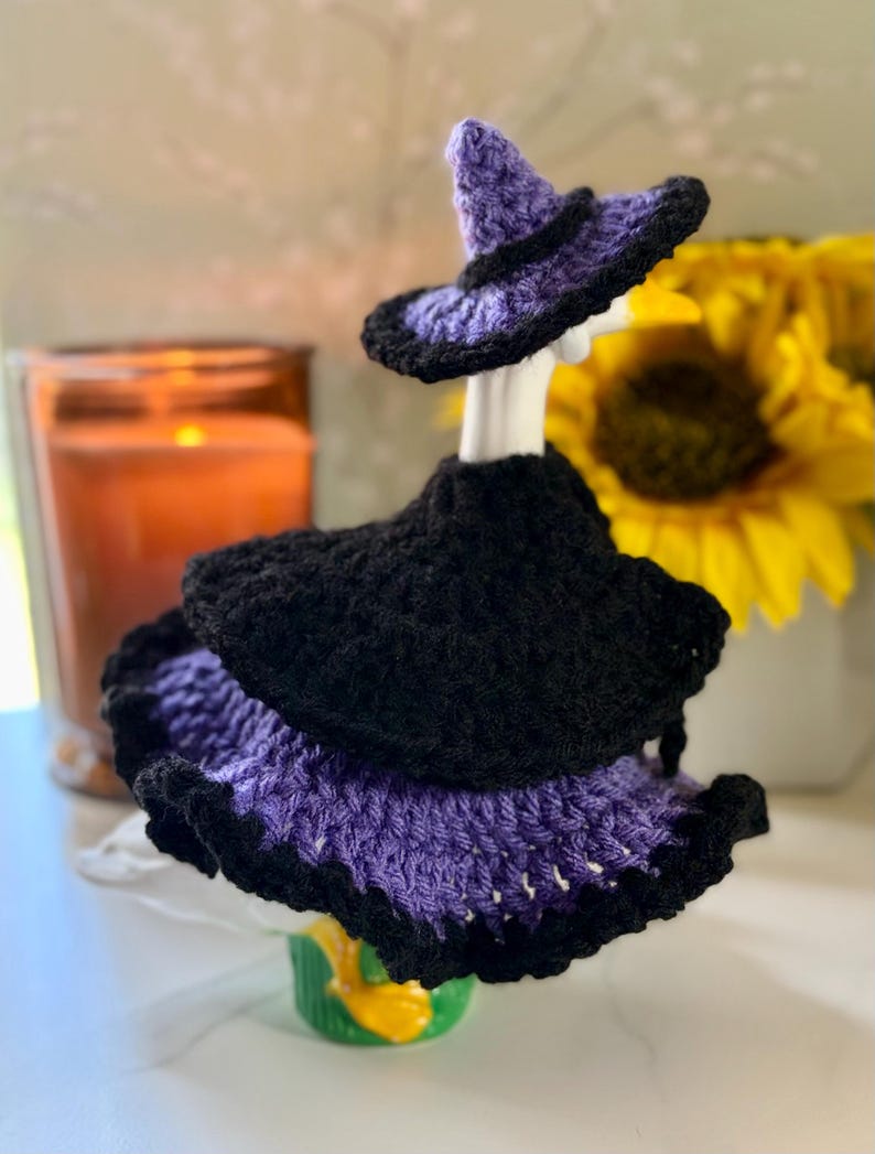 Pu&ograve; includere: Un completo all'uncinetto per una statuetta di oca composto da un cappello da strega nero e viola e un vestito a strati. Il vestito presenta un corpetto nero con una gonna viola e nera a balze. Il completo &egrave; progettato per un'oca bianca.