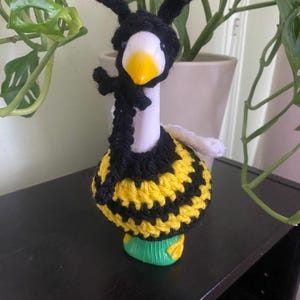 May include: A goose figurine in a bee costume. The goose has a white body and yellow beak, with a black and yellow crocheted bee outfit, including antennae and wings. The figurine is on a dark surface.