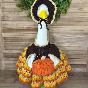 Pode incluir: Uma estatueta de ganso branco usando uma touca de crochê marrom escuro com acabamento creme e um vestido de crochê marrom escuro combinando com um padrão chevron em laranja, amarelo e bege. Uma pequena abóbora de crochê laranja está anexada.