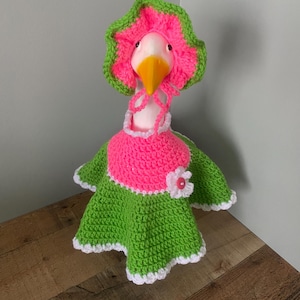 May include: A whimsical goose figurine wearing a handmade crochet outfit. The outfit includes a pink and green dress, a matching bonnet, and a white collar. The goose has a yellow beak and is set against a neutral background.