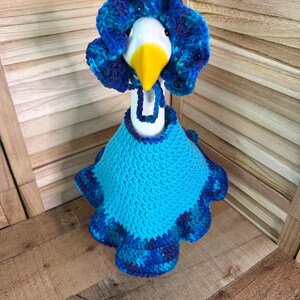 May include: A white ceramic goose figurine wearing a crocheted outfit consisting of a bright blue cape with a ruffled, variegated blue and purple border and a matching bonnet with a ruffled edge and a tie under the beak.