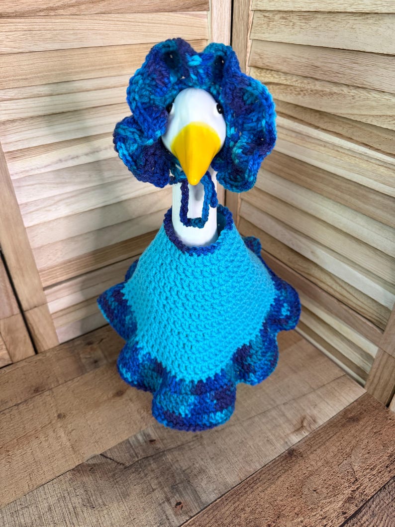 May include: A white ceramic goose figurine wearing a crocheted outfit consisting of a bright blue cape with a ruffled, variegated blue and purple border and a matching bonnet with a ruffled edge and a tie under the beak.