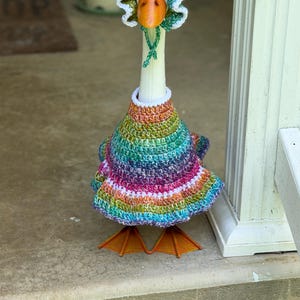May include: A white goose wearing a multi-colored, variegated yarn crochet dress with a white trim and a matching crochet bonnet featuring a ruffled edge and green ties. The dress displays a spectrum of rainbow hues including pink, blue, and green.