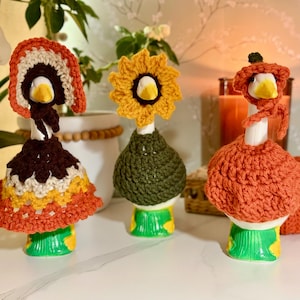May include: Three white ceramic goose figurines wearing handmade crochet outfits. The left goose wears a bonnet and a multi-colored dress. The center goose wears a yellow sunflower headpiece and a green sweater. The right goose wears an orange hat.