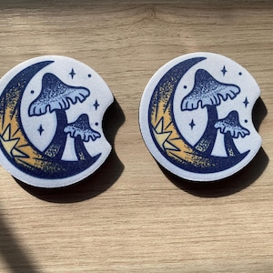 May include: Two car coasters with a whimsical design. Each coaster features a blue mushroom illustration set against a crescent moon with yellow and white accents. The background is white with small star details.