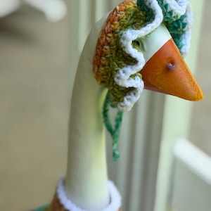 May include: A decorative goose figurine wearing a hand-crocheted bonnet and matching sweater. The bonnet features a ruffled white edge with variegated yarn in shades of green, orange, and yellow. The sweater has a white collar and a colorful pattern.