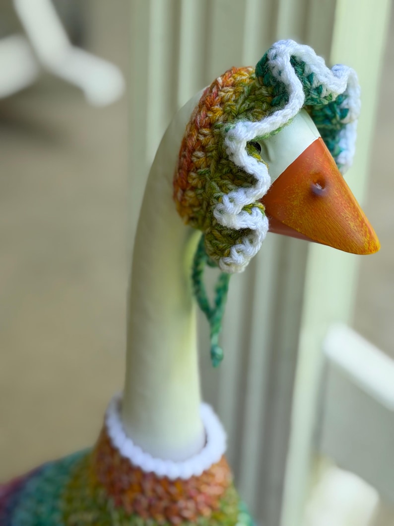 May include: A decorative goose figurine wearing a hand-crocheted bonnet and matching sweater. The bonnet features a ruffled white edge with variegated yarn in shades of green, orange, and yellow. The sweater has a white collar and a colorful pattern.