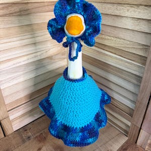 May include: A white goose figurine wearing a crocheted outfit consisting of a bright blue cape with a ruffled dark blue and purple variegated hem and a matching ruffled bonnet with ties, featuring a vibrant orange beak on the goose head.