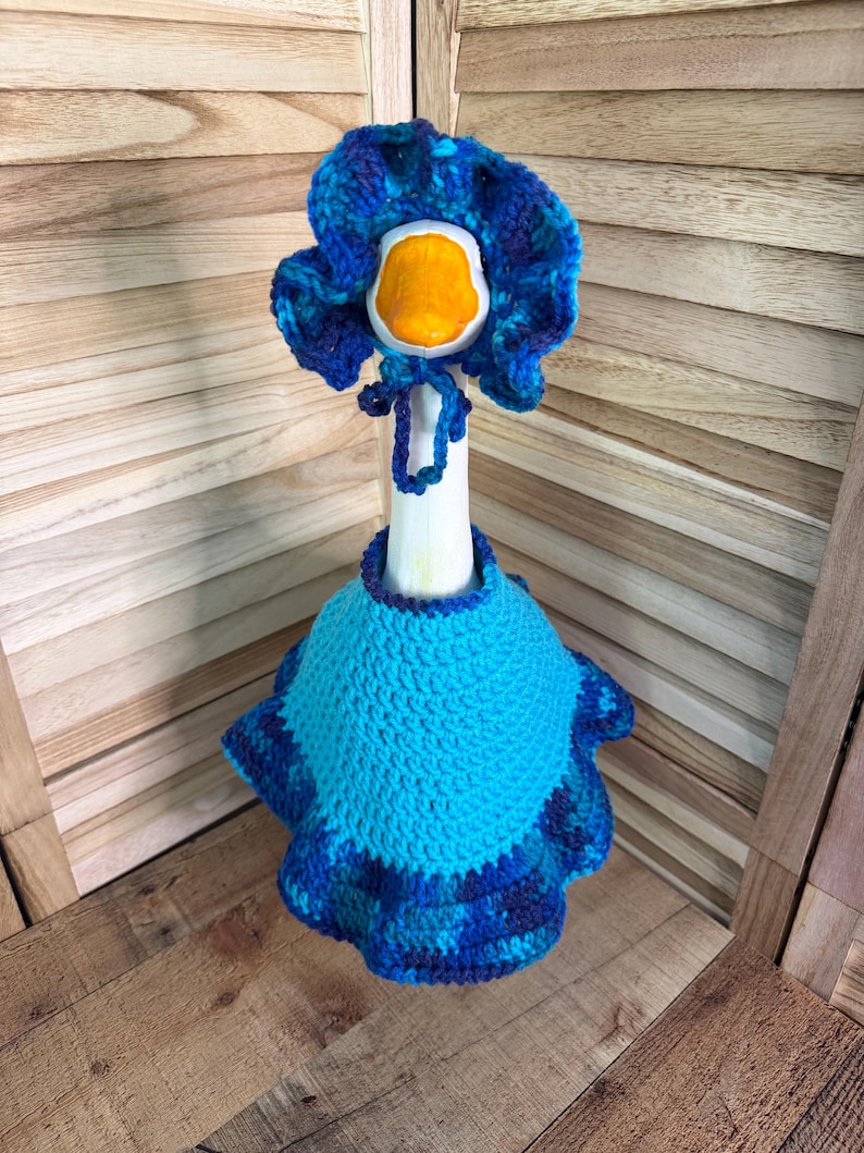 May include: A white goose figurine wearing a crocheted outfit consisting of a bright blue cape with a ruffled dark blue and purple variegated hem and a matching ruffled bonnet with ties, featuring a vibrant orange beak on the goose head.