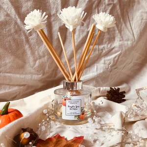 May include: A glass reed diffuser with a silver collar, containing liquid and white flower reeds. The label reads "Pumpkin Spice Reed Diffuser". The background features a pumpkin, pinecone, and fairy lights, creating a seasonal display.
