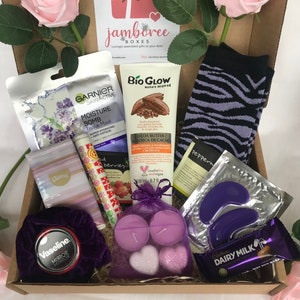 May include: A brown cardboard box filled with a variety of self-care items, including a face mask, body lotion, socks, eye masks, candles, and chocolate. The box is decorated with a pink rose and the text "jamboree boxes".