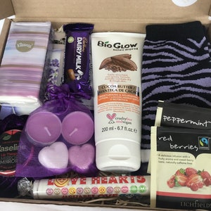 May include: A gift box filled with various items, including a purple and white patterned tissue box, a chocolate bar, a purple and black striped sock, a bottle of cocoa butter lotion, two purple heart-shaped candles, a small round tin with a purple lid, a small bag of peppermint tea, a small bag of red berries tea, and a tube of lip balm with the words "LOVE HEARTS" printed on it.