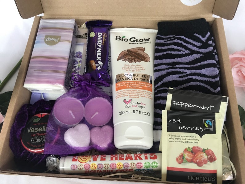May include: A gift box filled with various items, including a purple and white patterned tissue box, a chocolate bar, a purple and black striped sock, a bottle of cocoa butter lotion, two purple heart-shaped candles, a small round tin with a purple lid, a small bag of peppermint tea, a small bag of red berries tea, and a tube of lip balm with the words "LOVE HEARTS" printed on it.