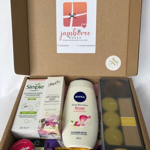 May include: A gift box filled with various items, including a bottle of rose-scented shower cream, a tube of hand cream, a bottle of body lotion, a small jar of lip balm, a bar of chocolate, and a pack of candles. The box is decorated with a thank you sticker and a logo for "jamboree boxes".