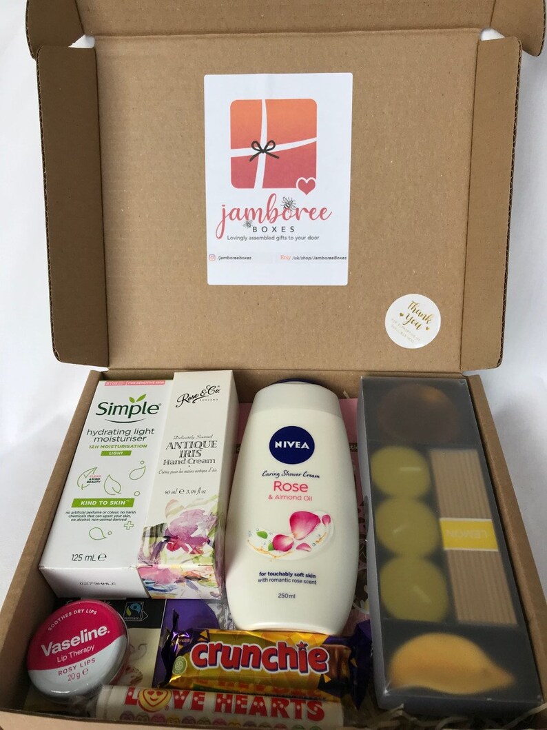May include: A gift box filled with various items, including a bottle of rose-scented shower cream, a tube of hand cream, a bottle of body lotion, a small jar of lip balm, a bar of chocolate, and a pack of candles. The box is decorated with a thank you sticker and a logo for "jamboree boxes".